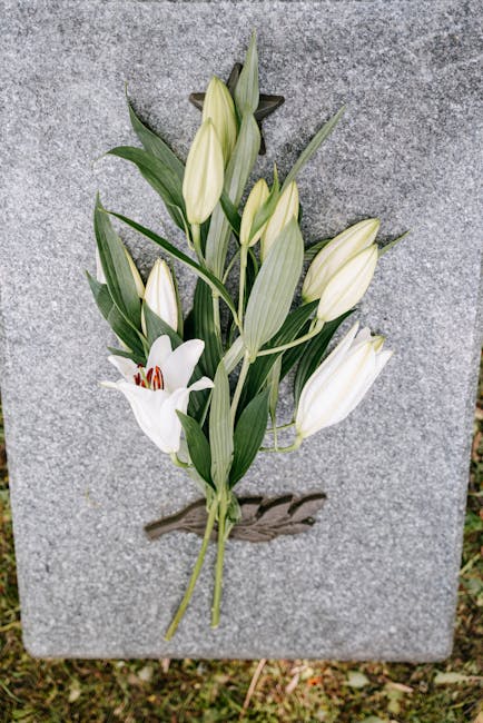 A white lily and lily buds with long, green leaves are arranged on a rectangular grey stone memorial. The lilies are fresh, with smooth petals and prominent stamens, and are laid neatly atop the stone