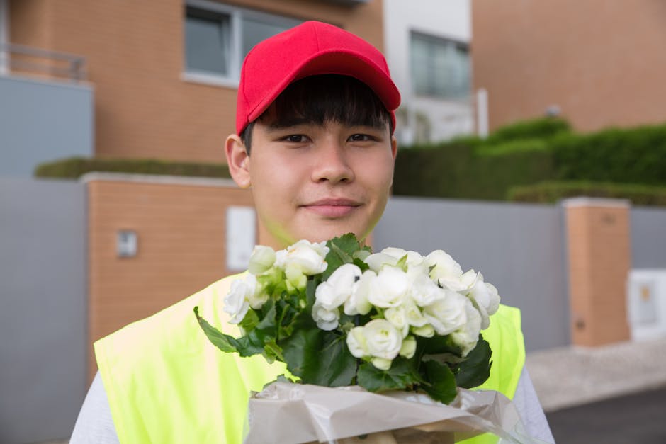 A young male delivery person wearing a red cap and a neon yellow safety vest is standing outdoors in front of modern residential buildings, holding a bouquet of fresh white flowers wrapped in white pa