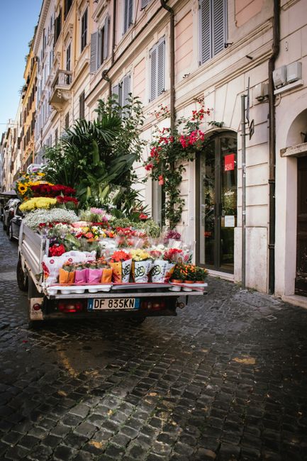 A flower delivery truck parked on a cobblestone street in front of a building with cream-colored walls and shuttered windows. The truck's flatbed is filled with vibrant, fresh floral arrangements, inc