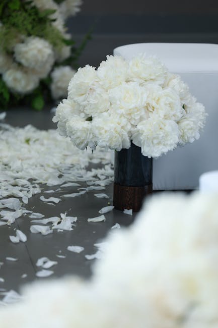 A round floral arrangement of white carnations placed in a tall, dark-colored vase with a wooden base, positioned on a table scattered with white flower petals, creating an elegant and fresh floral di