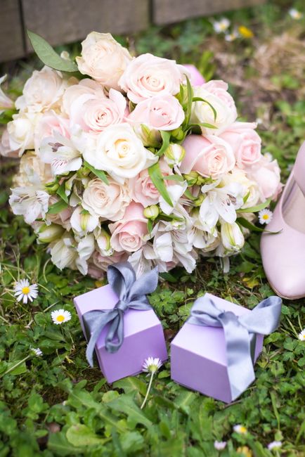 A floral arrangement comprising a round bouquet of fresh, pastel pink and white roses, interspersed with delicate white lilies and green foliage, displayed on the grass outdoors. The bouquet is wrappe